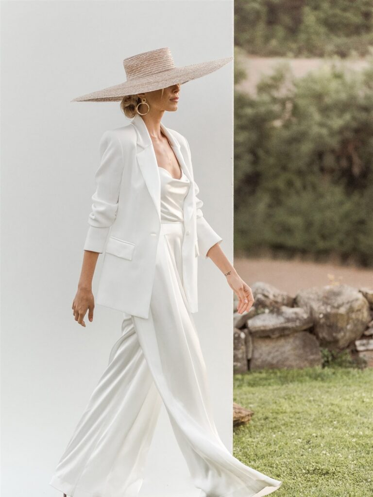 Model from the side in cool ivory white wedding suit. Wide-leg pants with high waist, draped satin top in ivory with spaghetti straps and slightly oversized jacket with rolled up long sleeves. Model wears large wicker hat.