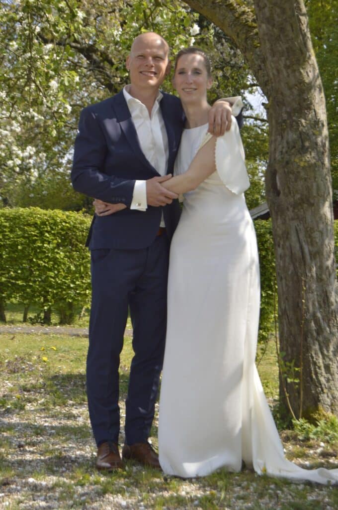 True Pippi bride Steffi with her husband. Steffi wears an ivory tight long wedding gown with butterfly sleeves that are cut on and fall open. The sleeves and high neckline are finished with a small pearl trim